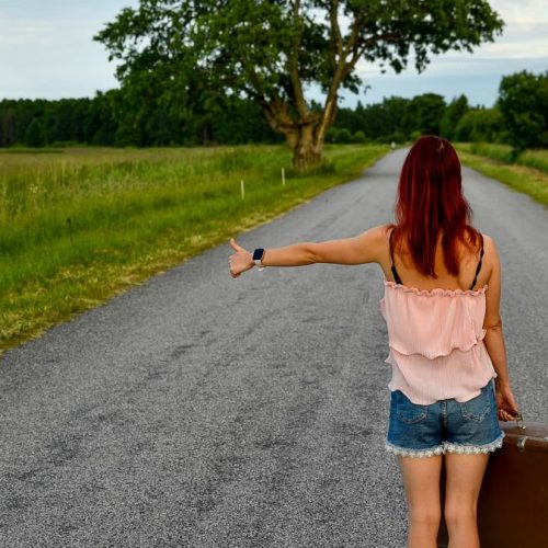 Woman hitchhiking on a rural road in Vihasoo, Estonia, carrying vintage luggage.