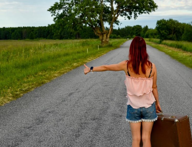 Woman hitchhiking on a rural road in Vihasoo, Estonia, carrying vintage luggage.