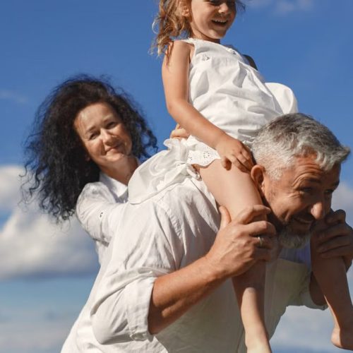 Grandparents playing with granddaughter outdoors under bright blue sky.