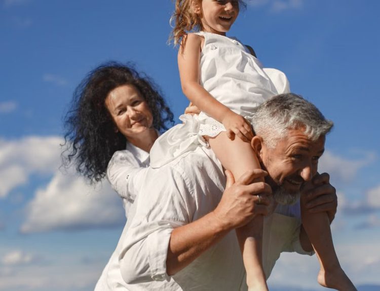 Grandparents playing with granddaughter outdoors under bright blue sky.