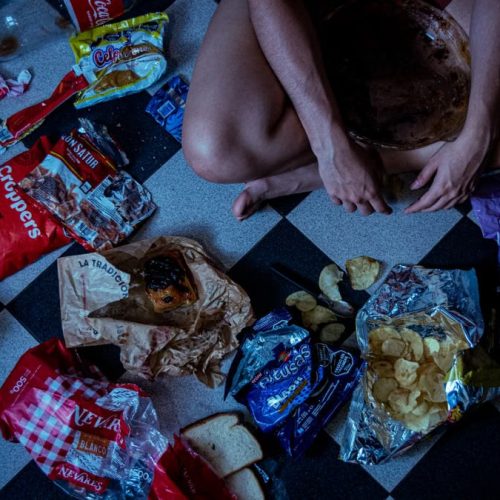 Man sitting surrounded by assorted snacks and packaging on a checkered floor, depicting a binge-eating scene.