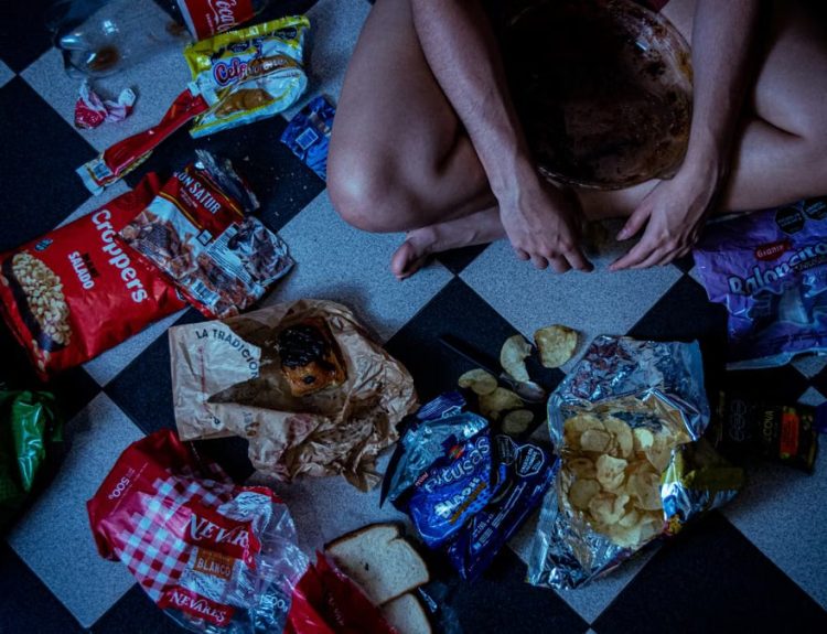Man sitting surrounded by assorted snacks and packaging on a checkered floor, depicting a binge-eating scene.